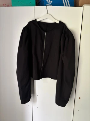 Svart cropped cardigan med puffärm - Stilren svart cardiganjacka med dragkedja framtill och trendiga puffärmar. Modellen är cropped och har rund halsringning. Materialet ser ut att vara ett mjukt tyg, perfekt för en cool och enkel look.