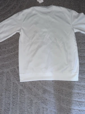 Vit sweatshirt från Lacoste - Klassisk vit sweatshirt från Lacoste med rund halsringning och den ikoniska krokodilloggan broderad på bröstet. Tröjan har lång ärm och ribbade muddar vid ärmslut och nederkant. Perfekt för en clean och stilren look.