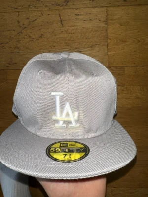 Grå LA New Era 59Fifty keps - Snygg grå fitted keps från New Era med LA-logga broderad framtill och MLB-logga bak. Klassisk 59Fifty-modell med rak skärm och strukturerad passform. Tillverkad i bomullstyg som ger en clean och stilren look.