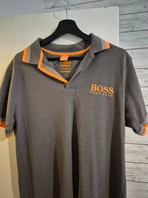 Grå pikétröja från Hugo Boss - Snygg grå pikétröja från Hugo Boss med orange detaljer på krage och ärmslut. Tröjan har korta ärmar och klassisk krage med orange logga på bröstet. Perfekt för en stilren och sportig look.