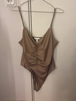 Beige body med rynk framtill H&M - Säljer en beige body från H&M med smala axelband och snygg rynkdetalj framtill. Modellen har djup ringning och är tillverkad i ett mjukt, stretchigt material som sitter skönt mot kroppen. Perfekt att styla med jeans eller kjol.