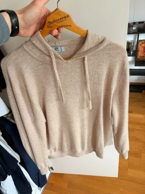 BVALDI Hoodie - Mysig beige hoodie från BVALDI med broderad logga på bröstet. Tillverkad i mjukt stickat material och har dragsko i huvan. Perfekt för en chill och avslappnad stil. Långa ärmar och ribbade muddar för extra komfort.