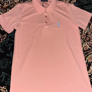 Rosa pikétröja från Polo Ralph Lauren - Snygg rosa pikétröja från Polo Ralph Lauren med klassisk krage och knappar framtill. Tröjan har det ikoniska blå broderade logotypen på bröstet och är tillverkad i mjuk bomull. Perfekt för en avslappnad men stilren look.
