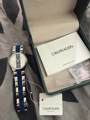 Calvin Klein blå och silver klocka - Snygg klocka från Calvin Klein med blått och silverfärgat armband i metall och syntet. Knappt använt. Men boxen till Calvin Klein den gick sönder så hittade en annan som passar. Men har knappt använt den och den är jättefin