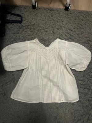 Vit romantisk blus med puffärm - Söt vit blus med breda puffärmar och spetsdetaljer. Blusen har dekorativa sömmar framtill samt fina veck och spets längs ärmar och bröst. Tillverkad i ett tunt, luftigt tyg som ger en bohemisk känsla. Den är aldrig använd. Nypris: 450
