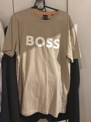 Beige BOSS t-shirt med tryck - Snygg beige t-shirt från BOSS med stort vitt logotryck på bröstet. Klassisk rund halsringning och korta ärmar. Tillverkad i mjuk bomull för en skön känsla och avslappnad passform.