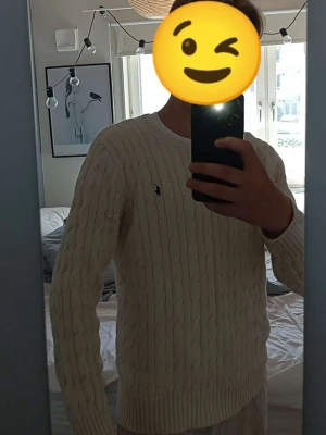 Vit kabelstickad tröja från Polo Ralph Lauren - Säljer en klassisk vit kabelstickad tröja från Polo Ralph Lauren med rund halsringning och den ikoniska broderade loggan på bröstet. Tröjan har långa ärmar och ribbade muddar vid ärmslut och nederkant. Perfekt för en stilren och tidlös look. Storlek L (14-16) sitter bra för 175cm +