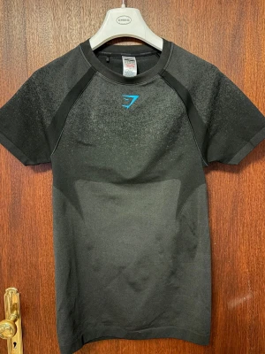 Gymshark tröja - Snygg svart träningströja från Gymshark med korta ärmar och blå logga på bröstet. Tillverkad i ett stretchigt funktionsmaterial som andas, perfekt för gymmet eller löpning. Tröjan har en tight passform och diskreta mönsterdetaljer på axlar och rygg.