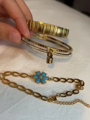 Guldarmband - Tre snygga armband i guldton: ett med graverade linjer, ett med gnistrande stenar och hängande lås, samt ett kedjearmband med en blå blomma av pärlor i mitten. Perfekta för att mixa och matcha till din stil.