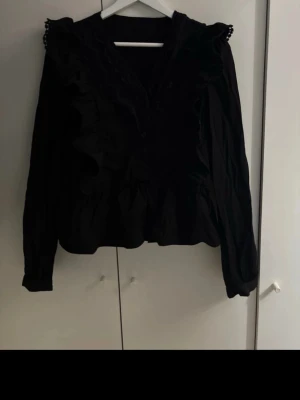 Svart volangblus med spetsdetaljer - Supersnygg svart blus med volanger längs axlar och bröst, samt spetsdetaljer vid halsringningen. Blusen har knappar framtill och en lätt rynkad midja som ger en feminin siluett. Perfekt till jeans eller kjol för en trendig look.