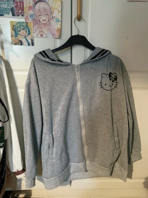 Grå Hello Kitty hoodie med dragkedja - Säljer en grå hoodie med dragkedja från Hello Kitty. På bröstet finns ett tryck av Hello Kitty med en svart rosett och dödskalle. Hoodien har huva med svarta ränder och två fickor framtill. Köpt på Shopee och använd en gång. Utan defekter eller skador. Original pris ca ~250kr
