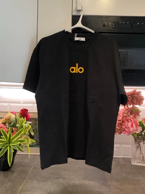 Svart t-shirt från Alo med gul text - Snygg svart t-shirt från Alo med gul broderad text på bröstet. Klassisk rund halsringning och korta ärmar. Tillverkad i mjuk bomull som känns skön mot huden. Perfekt för en clean och enkel stil.