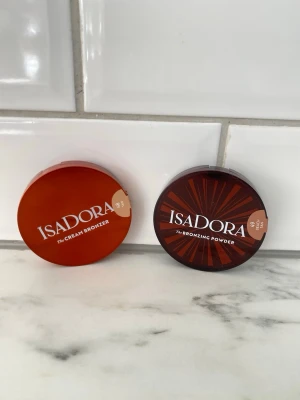 Isadora bronzer duo i bruntoner - Säljer två bronzers från Isadora: en The Cream Bronzer och en The Bronzing Powder. Båda kommer i runda, bruna plastförpackningar med tydlig logga. Perfekta för att skapa en solkysst look och ge ansiktet värme och lyster.