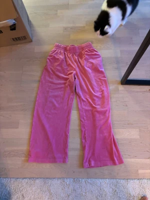 Rosa velourbyxor med resårmidja - Mysiga loungewear-byxor i rosa velour med bred resår i midjan och raka ben. Perfekta för chill hemma eller när du vill vara extra bekväm. Materialet är mjukt och stretchigt för maximal komfort.