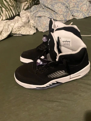 Svarta Air Jordan 5 Retro sneakers - Snygga Air Jordan 5 Retro sneakers i svart mocka med vita och grå detaljer. Skorna har den ikoniska Jumpman-loggan på plösen, genomskinliga snörlås och meshpaneler på sidorna. Tjock vit sula med synlig luftbubbla och coola detaljer längs sidan.