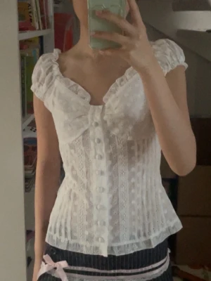 Liknar brandy melville, Vit blus med spets och rosett - Liknar brandy melville. Söt vit blus med puffärmar, spetsdetaljer och stor rosett framtill. Blusen har knappar längs hela framsidan och ett figurnära snitt. Materialet är tunt och har broderade mönster som ger en romantisk känsla. Perfekt för dig som gillar vintage eller cottagecore-stil.