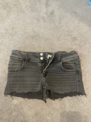 Svarta jeansshorts  - Säljer ett par svarta jeansshorts med dubbla knappar i midjan. Shortsen har klassisk femficksdesign och är perfekta för en avslappnad stil. Passar dig som gillar en edgy look.