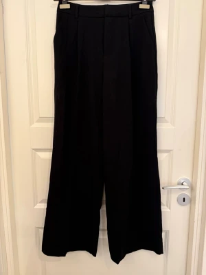 Svarta vida kostymbyxor - Säljer ett par svarta kostymbyxor med hög midja och vida ben. Byxorna har pressveck framtill, bälteshällor och sidofickor. Perfekta för en clean och stilren look. Materialet ser ut att vara en mjuk och följsam textil. Oanvänd med prislapp. 