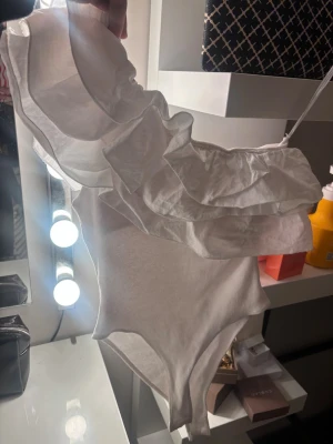 Vit body med volang från Zara - Supersnygg vit body från Zara med stora volanger över axeln och ena sidan. Tillverkad i ribbat tyg som ger en skön och stretchig känsla. Perfekt för dig som vill ha något extra till outfiten. Axelbandslös design som ger en cool och trendig look. Super fin, inga fläckar eller sådant.