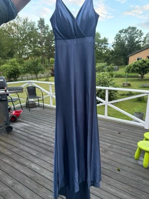 Marinblå långklänning i satin - Elegant marinblå långklänning i glansigt satinmaterial med smala axelband och v-ringad omlottringning. Klänningen har en figurnära passform upptill och faller mjukt längs kroppen. Perfekt för speciella tillfällen.