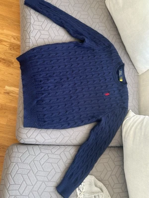 Mörkblå kabelstickad tröja Polo Ralph Lauren - Storlek L men passar M med. Kabelstickad mörkblå tröja från Polo Ralph Lauren med rund halsringning och den klassiska röda logotypen broderad på bröstet. Tröjan har långa ärmar och ribbade muddar vid ärmslut och nederkant. Perfekt för en stilren och tidlös look. 