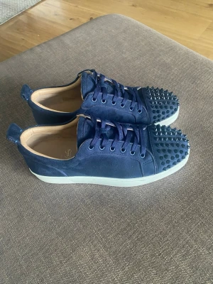 Blåa sneakers med nitar från Louboutin - Snygga mörkblå sneakers från Christian Louboutin med matta nitar på tån, vita sulor och blå mocka. Skorna har klassisk låg profil, rund tå och matchande blå skosnören. Perfekta för dig som vill sticka ut med en edgy detalj. Storlek 42, kan även passa som 42