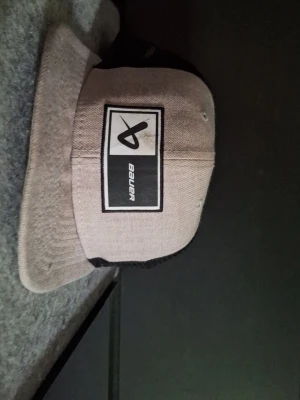 Grå keps från Bauer med logga - Snygg beige keps från Bauer med svart och vit rektangulär logga framtill. Kepsen har platt skärm och mesh på baksidan för extra ventilation. Perfekt för dig som gillar sportig och avslappnad stil.