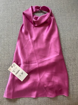 Rosa halterneck linnetopp i satin - Trendig rosa halterneck topp från Zara i glansigt satinmaterial. Toppen har en snygg draperad halsringning och är ärmlös med öppen rygg. Perfekt för dig som vill sticka ut med en färgstark och elegant look.