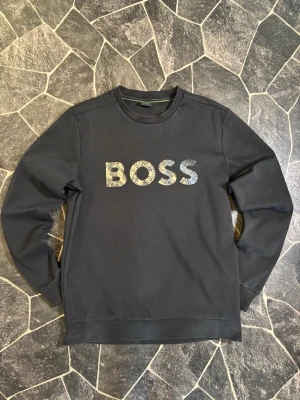 Boss Sweatshirt  - Stilren svart sweatshirt från BOSS med rund halsringning och stort BOSS-tryck i grått framtill. Tröjan har långa ärmar och ribbade muddar vid ärmslut och nederkant. Perfekt för en avslappnad och trendig look.