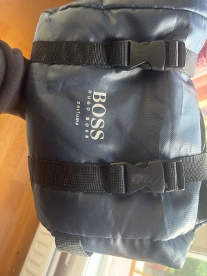 Marinblå shoppingväska från Hugo Boss - Snygg marinblå shoppingväska från Hugo Boss Parfums med två svarta spännen framtill. Väskan är tillverkad i slitstark nylon och har en enkel, sportig design med tryckt logga på framsidan. Perfekt för dig som vill ha en praktisk och stilren väska.