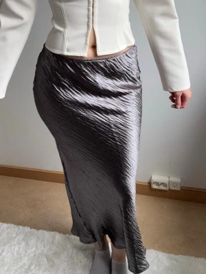 satin kjol  - Säljer en glansig silvrig långkjol med snyggt vågig struktur. Kjolen har en figurnära passform och faller mjukt längs benen. Perfekt för dig som vill ha en stilren och trendig look. Inga fickor eller synliga detaljer.