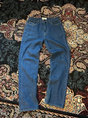 Blå raka jeans  - Säljer ett par klassiska blå jeans med rak passform och hög midja. Jeansen har fem fickor, bälteshällor och stängs med dragkedja och knapp. Materialet är denim i 100% bomull för en tidlös look. Storlekenen är 30-32