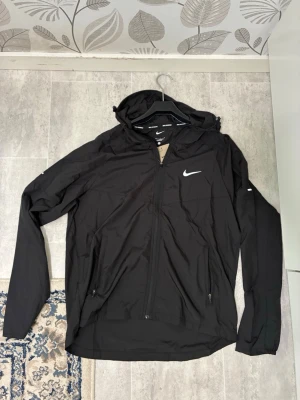 Svart vindjacka från Nike Miller - Snygg svart vindjacka från Nike Running med huva och dragkedja framtill. Jackan har två fickor med dragkedja, justerbar huva och vit Nike-logga på bröstet. Perfekt för löprundan eller när du vill ha en sportig look.