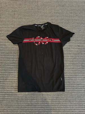 Svart t-shirt med ormtryck från Black Squad - Svart t-shirt från Black Squad med ett iögonfallande tryck av en röd, vit och svart orm över gröna och röda ränder på bröstet. Klassisk rund hals och korta ärmar. Perfekt för dig som vill sticka ut med en cool detalj.