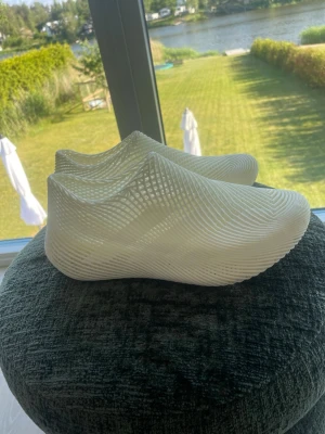 Vita 3D-printade tofflor - Säljer ett par vita tofflor med futuristisk 3D-printad design. De har en slät, vågig struktur och är tillverkade i ett plastliknande material. Perfekta för dig som gillar unika och moderna skor med en clean look.