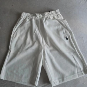 Vita shorts från Polo Ralph Lauren - Säljer ett par klassiska vita shorts från Polo Ralph Lauren med elastisk midja och sidofickor. Shortsen har den ikoniska loggan broderad på vänster ben och är tillverkade i mjukt bomullsmaterial för en skön känsla.