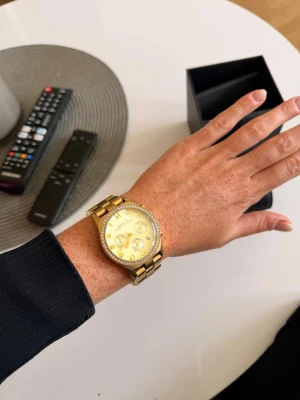 Guldig klocka från Marc Jacobs - Snygg guldig klocka från Marc Jacobs med länkarmband i metall och glittrande stenar runt urtavlan. Urtavlan har flera små visare och tydliga markeringar. Perfekt accessoar för att lyfta din stil. Batteriet på klockan behöver bytas!