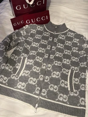  Gucci - Snygg grå stickad ziptröja från Gucci med vit GG-monogram över hela plagget. Tröjan har hög krage, två fickor framtill och ribbade muddar. Perfekt för dig som vill ha en exklusiv och trendig look.