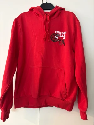 Röd Chicago Bulls hoodie med huva - Snygg röd hoodie med Chicago Bulls-logga på bröstet. Klassisk känguruficka framtill, ribbade muddar och justerbar huva med snören. Perfekt för dig som gillar basket och streetwear. Materialet känns mjukt och bekvämt, passar till chill vardagsstil.