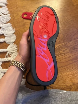 Christian Louboutin sneakers - Snygga sneakers från Christian Louboutin med den ikoniska röda sulan och svart ovandel. Skorna har en modern och stilren design med tydlig logotyp på sulan. Perfekta för dig som vill sticka ut med exklusiva detaljer och lyxig känsla.