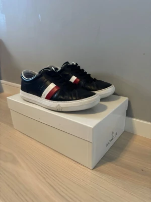 Moncler svarta sneakers med ränder - Snygga svarta sneakers från Moncler med vita och röda ränder på sidan samt vit sula. Skorna har klassisk låg profil, svarta skosnören och är tillverkade i läder. Perfekta för dig som gillar stilrena och exklusiva sneakers.