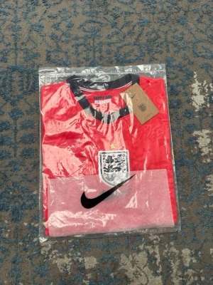 England Nike röd matchtröja - Säljer en röd England matchtröja från Nike med svart och vit detalj vid krage och ärmslut. Tröjan har England-emblemet på bröstet och ett stort svart Nike-märke. Tillverkad i lätt och ventilerande material, perfekt för fotboll eller träning.