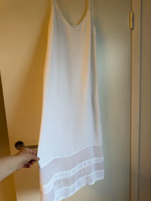Vit somrig klänning med tunna axelband - Säljer en enkel och stilren vit klänning med tunna axelband och v-ringning. Klänningen har transparenta detaljer längst ner och är gjord i ett lätt, svalt material som passar perfekt till varma dagar.