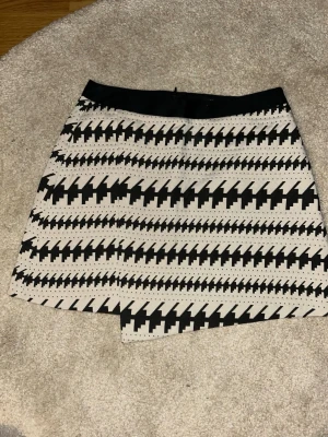 Svartvit kjol med grafiskt mönster - Säljer en svartvit kjol med coolt grafiskt zigzag-mönster. Skorten har en omlott-look framtill och bred midja. Materialet känns stadigt och har en lätt struktur. Perfekt för dig som vill sticka ut med en unik stil!