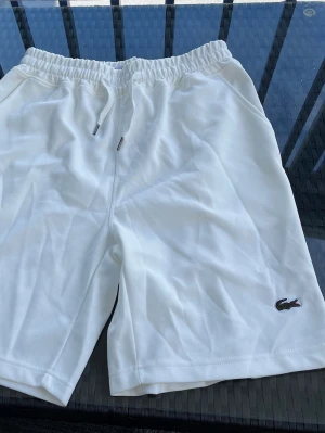 Vita shorts från Lacoste med snörning - Säljer ett par vita shorts från Lacoste med klassisk krokodillogga broderad på benet. Shortsen har elastisk midja med snörning och sidofickor. Perfekta för sommaren och riktigt clean look.