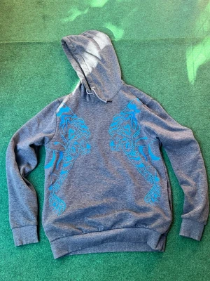 Grå hoodie med blå tigertryck - Snygg grå hoodie med huva och dragsko. På framsidan finns ett coolt blått tigerinspirerat tryck på båda sidor. Hoodien har ribbade muddar vid ärmslut och nederkant. Perfekt för dig som vill sticka ut med en unik design.