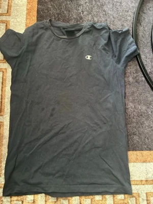 Svart Champion t-shirt i bomull - Säljer en svart t-shirt från Champion i mjuk bomull. Klassisk passform med korta ärmar och rund halsringning. Diskret Champion-logga på bröstet. Perfekt till jeans eller shorts för en enkel och clean look.