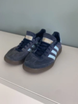 Adidas blå sneakers med vita ränder - Mörkblå sneakers från Adidas med klassiska vita ränder på sidorna och brun gummisula. Ovandelen är i mocka och skorna har blå snören. Perfekt för dig som gillar retrostil och streetwear.