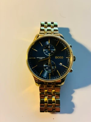 Guldfärgad klocka från BOSS - Stilren guldfärgad klocka från BOSS med blå urtavla och guldiga detaljer. Klockan har länkarmband i metall, tre subdials och datumvisning. Perfekt accessoar för dig som vill ha en klassisk men modern look.