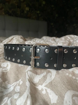 NYTT Svart nitskärp i konstläder - Snyggt svart bälte med silverfärgade nitar och spänne. Bältet är brett och i konstläder. Passar till jeans eller över en klänning för en cool look Längd: 105cm Från spännet till mittersta hålet är det 90cm